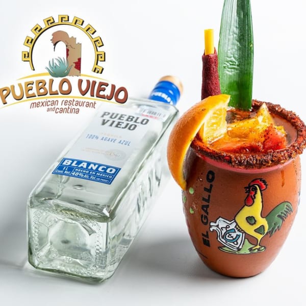 Pueblo Viejo Blanco tequila bottle next to a michelada in a clay mug with an orange slice.