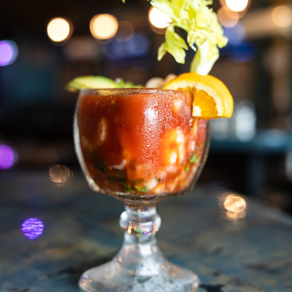 A vibrant red Mexican shrimp cocktail, garnished with celery and an orange slice, on a dark table.