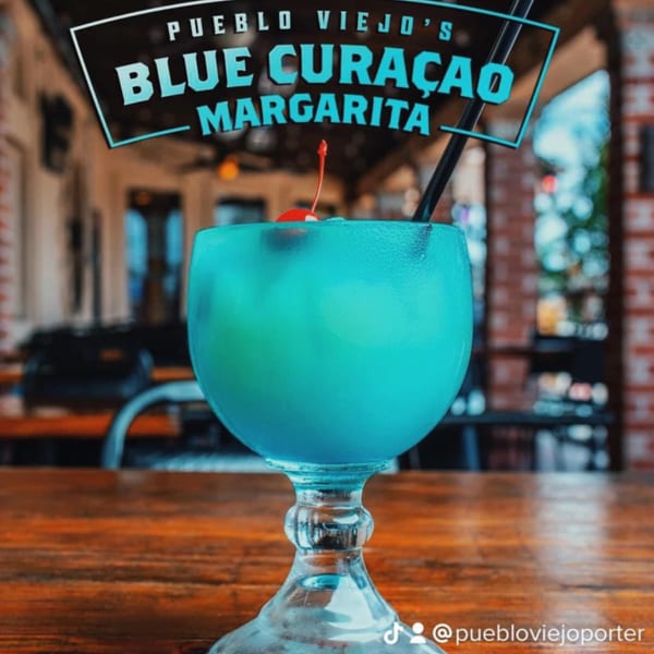Pueblo Viejo's Blue Curaçao Margarita, garnished with a cherry and straw, on a wooden table.