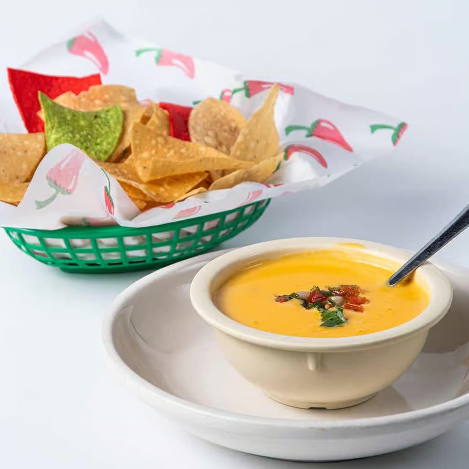 A bowl of yellow queso dip with a spoon, next to a basket of tortilla chips.