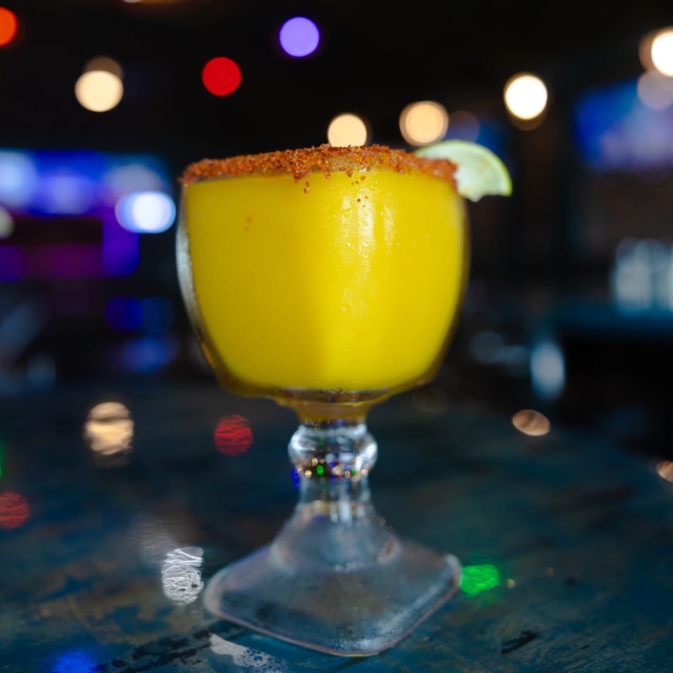 A vibrant yellow mango margarita with a chili-lime rim and lime wedge, against a blurred, colorful background.
