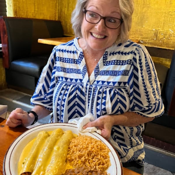 A smiling woman in glasses holds a plate of enchiladas, rice, and beans at Pueblo Viejo.
