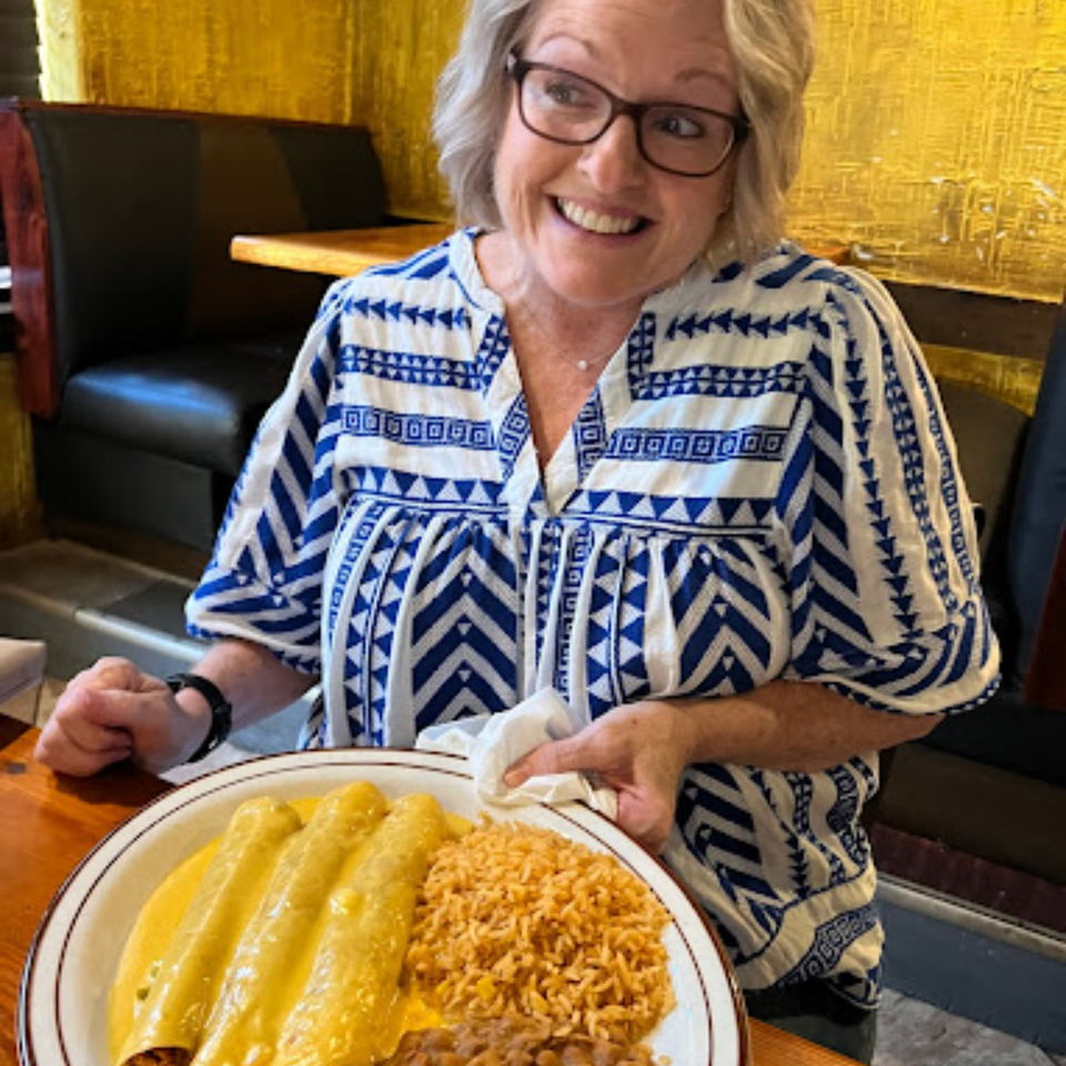 A smiling woman in glasses holds a plate of enchiladas, rice, and beans at Pueblo Viejo.