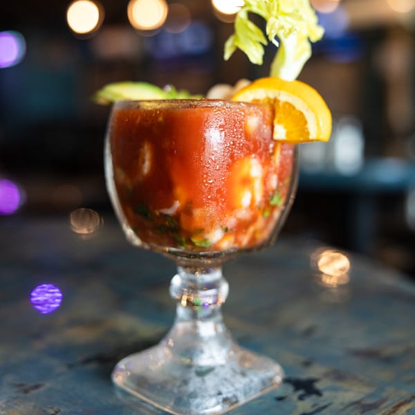 A vibrant red Michelada cocktail, garnished with celery and an orange slice, sits on a table.