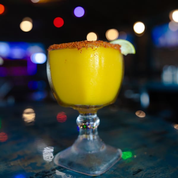 A vibrant yellow margarita with a chili-lime rim and lime wedge, against a blurred, colorful background.