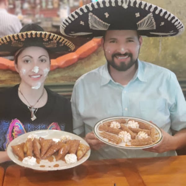 Two smiling people in sombreros with whipped cream on their faces, holding plates of Mexican desserts.