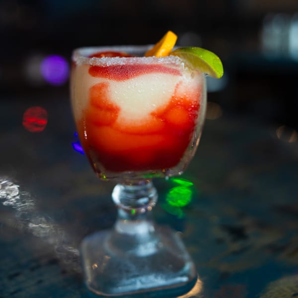 Strawberry margarita with a salted rim, garnished with lime and orange slices, on a dark, reflective surface.