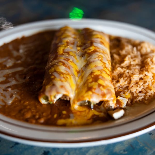 Two cheesy enchiladas covered in red sauce, served with refried beans and rice on a white plate.