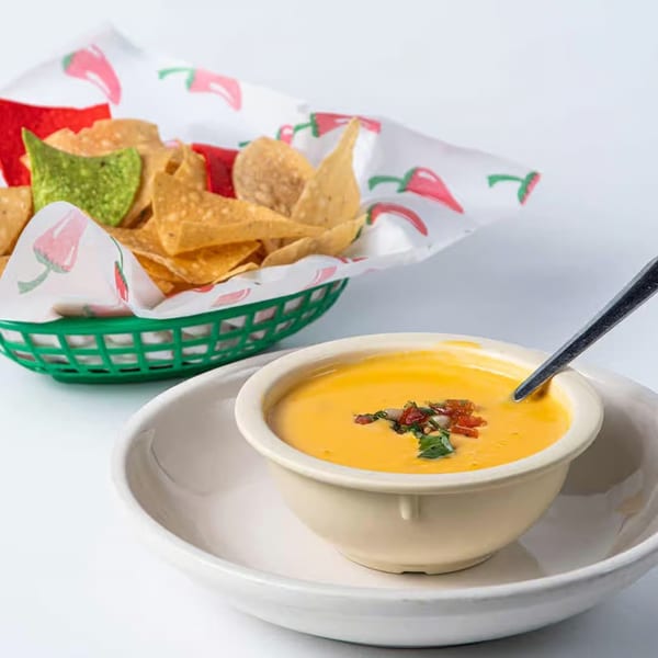 A bowl of yellow queso dip with red and green toppings, next to a basket of tortilla chips.
